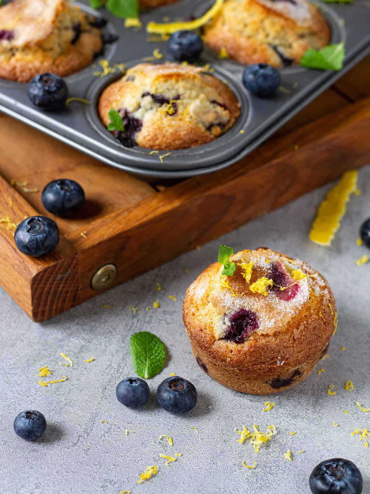 Freshly baked blueberry muffin cupcakes with lemon zest mint in metal baking tin and wooden tray on grey background