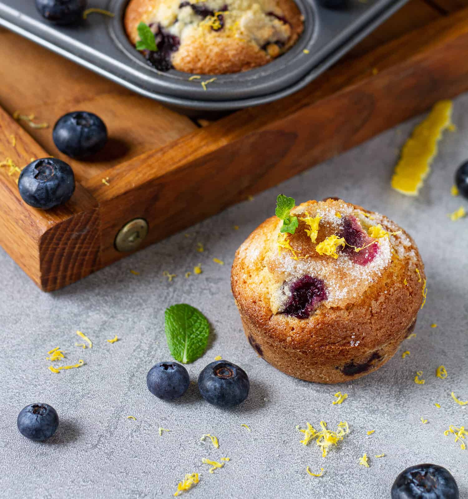 Freshly baked blueberry muffin cupcakes with lemon zest mint in metal baking tin and wooden tray on grey background 