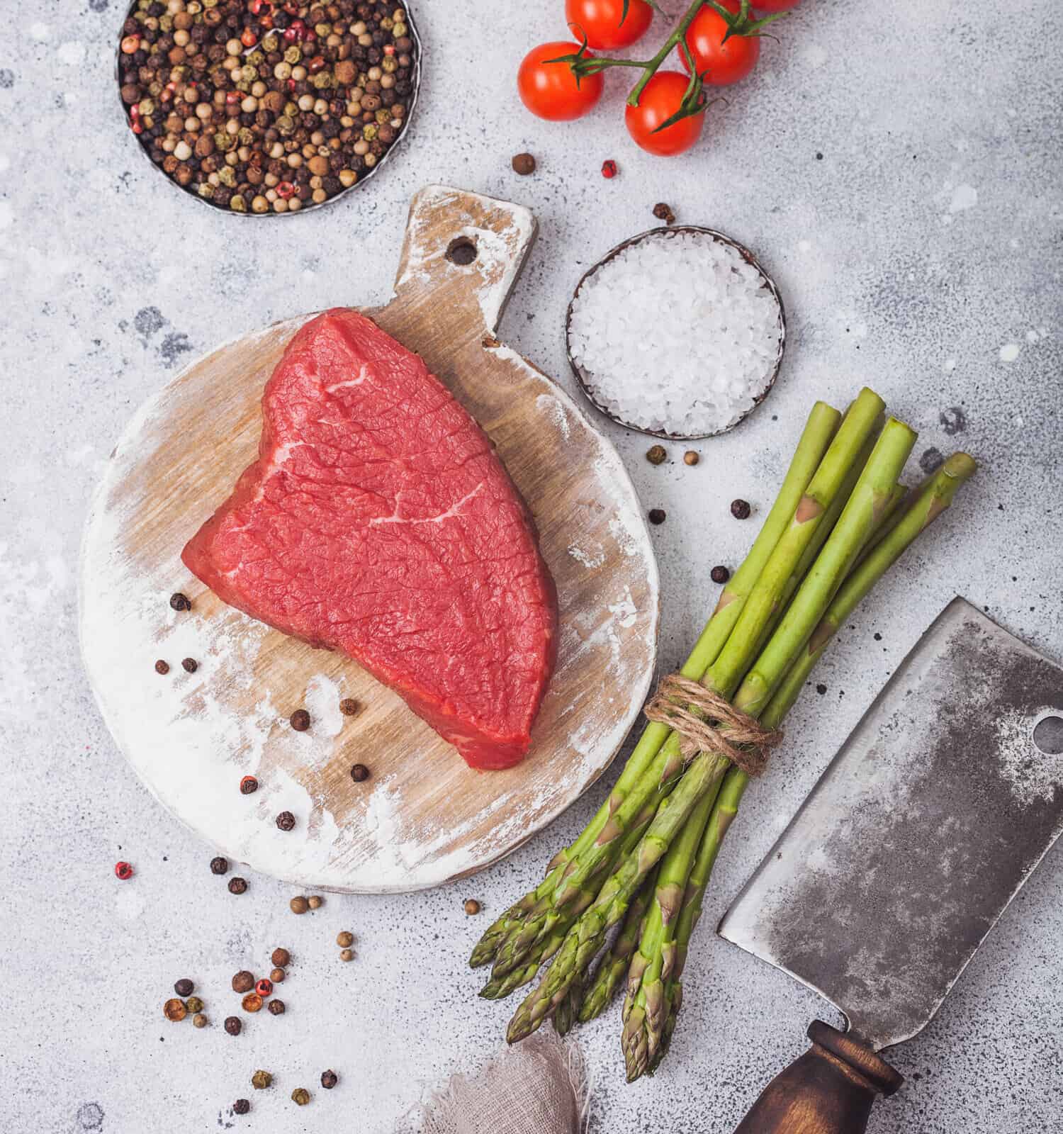 Slice of Raw Beef sirlion steak on round chopping board with tomatoes,garlic and asparagus tips and meat hatchet on light kitchen table background.