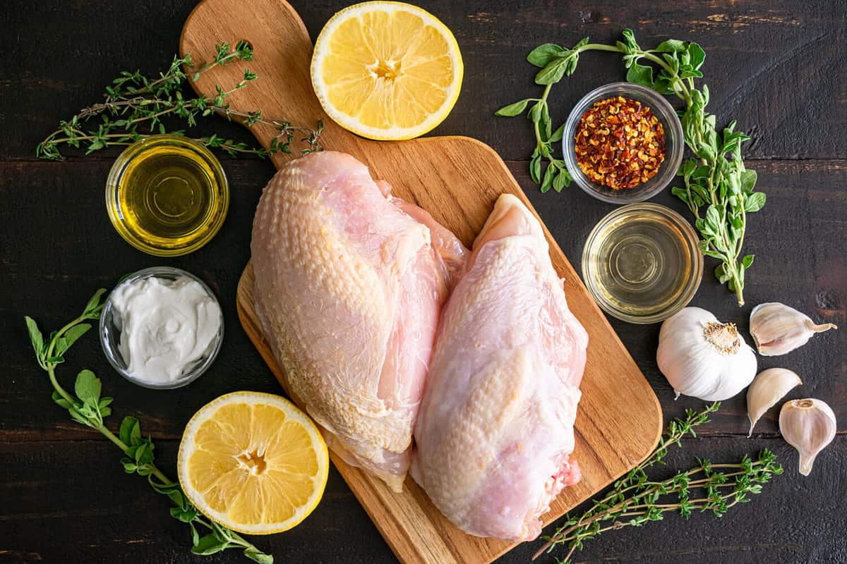 Oven-Roasted Greek Chicken Ingredients on a Dark Wood Background: Raw chicken breasts, lemon, herbs, and other ingredients