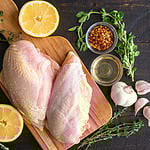Oven-Roasted Greek Chicken Ingredients on a Dark Wood Background: Raw chicken breasts, lemon, herbs, and other ingredients