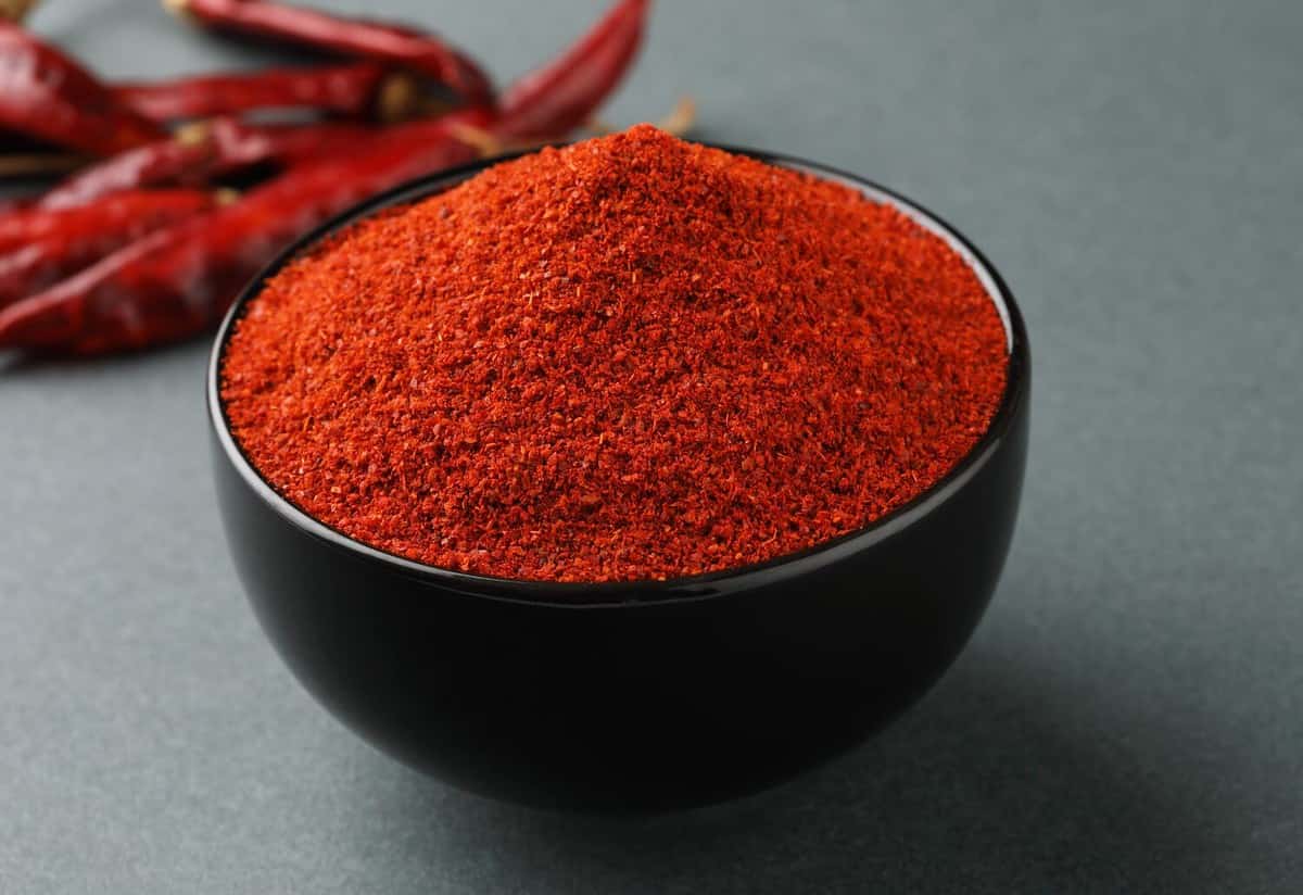 Chili powder is a seasoning blend of ground dried chiles in a black bowl with dried red chiles on a gray background.