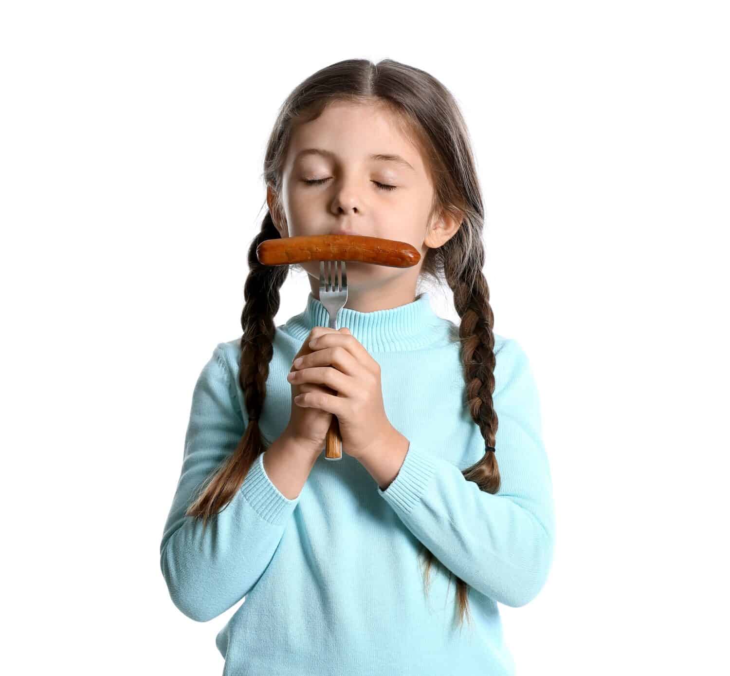 Cute little girl eating tasty sausage on white background
