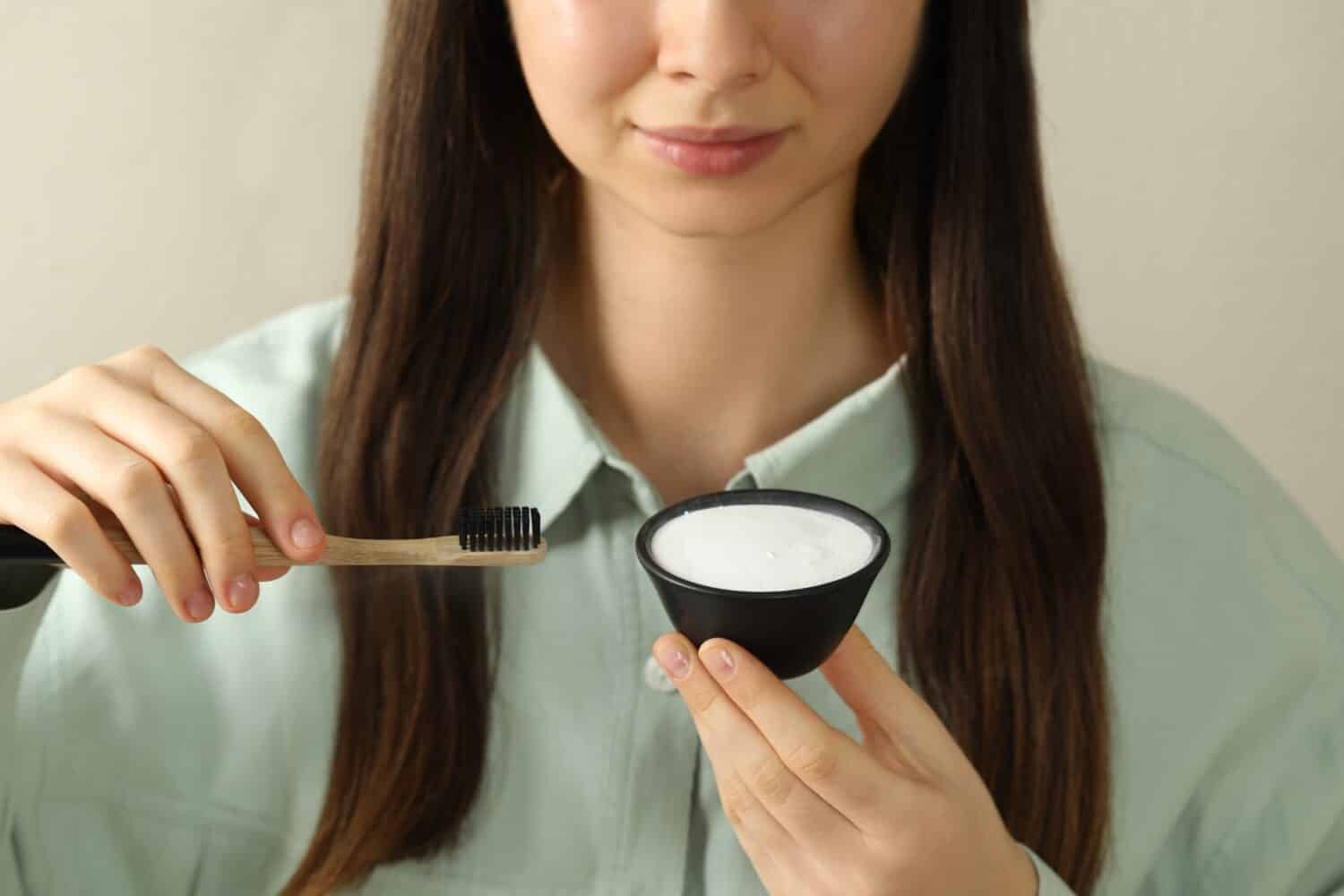 Young woman with toothbrush and bowl of baking soda on beige background, closeup
