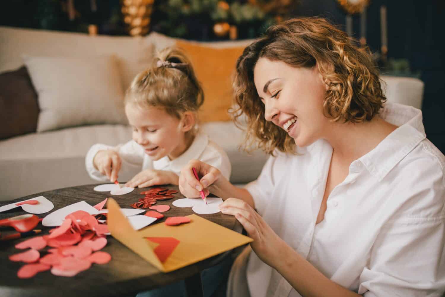 Mother and little daughter making Valentine's day cards using color paper, scissors and pencil, sitting by the table in cozy room