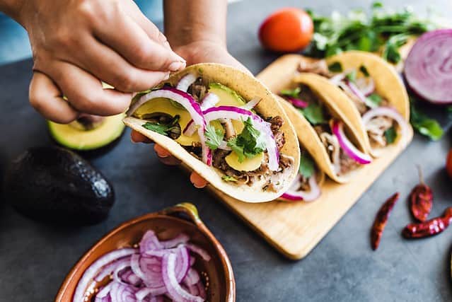 latin woman hands preparing mexican tacos with pork carnitas, avocado, onion, cilantro, and red sauce in Mexico