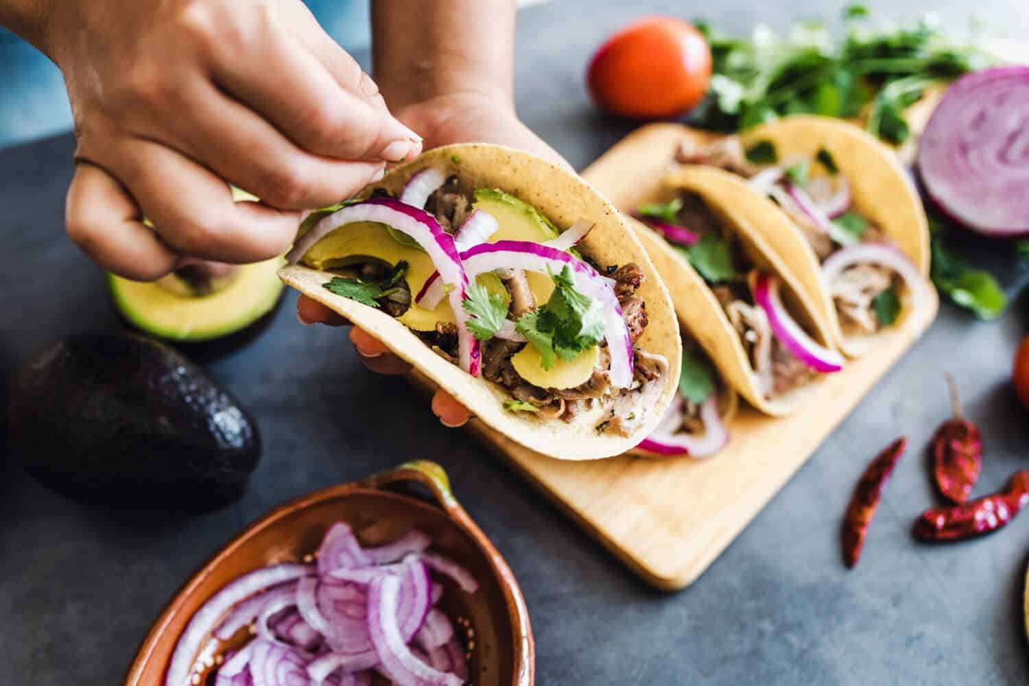 latin woman hands preparing mexican tacos with pork carnitas, avocado, onion, cilantro, and red sauce in Mexico