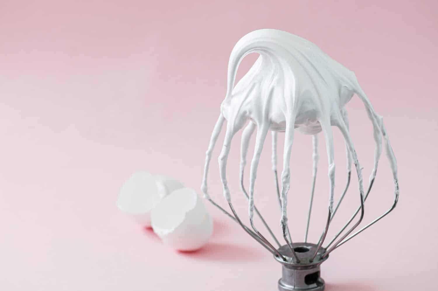 Whipped egg whites - beaten italian meringue on a wire whisk and egg shells on pink background. 