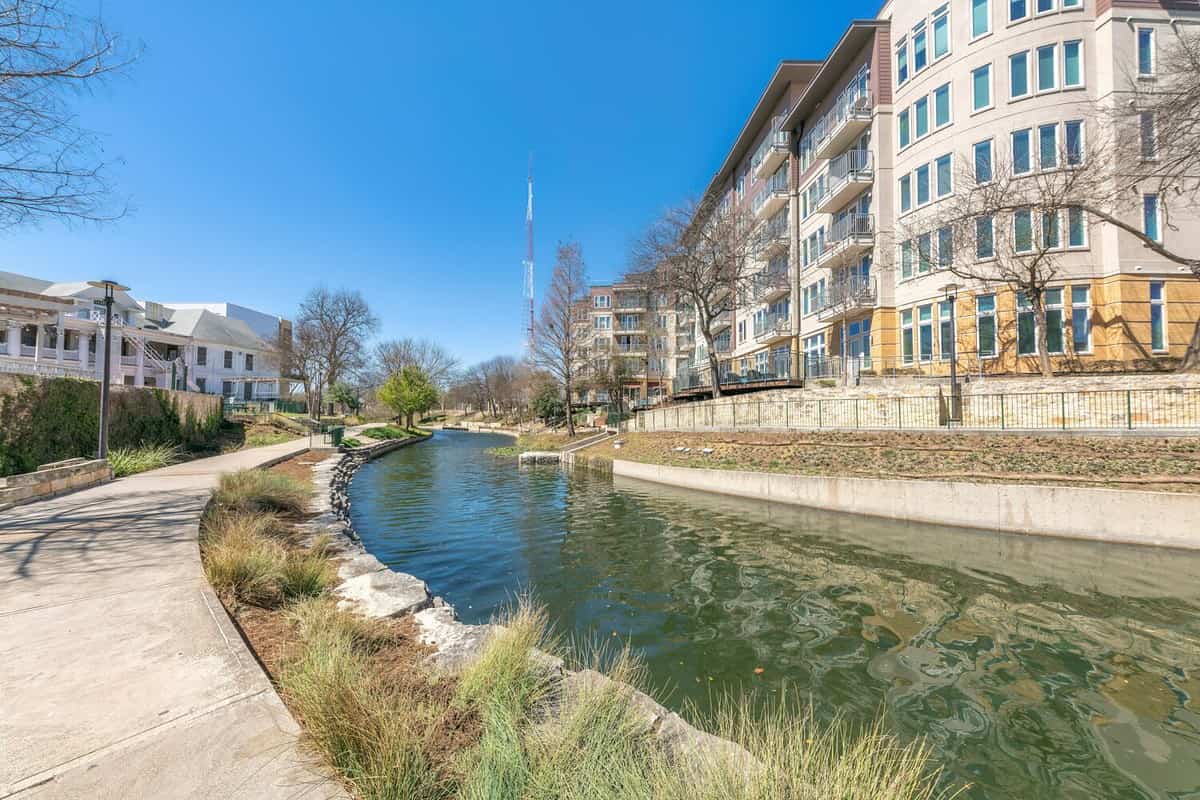Small river in the middle of mid-rise residential buildings at San Antonio, TX. There is a concrete walkway on the left side of the river and view of a tower at the back of the building on the right.