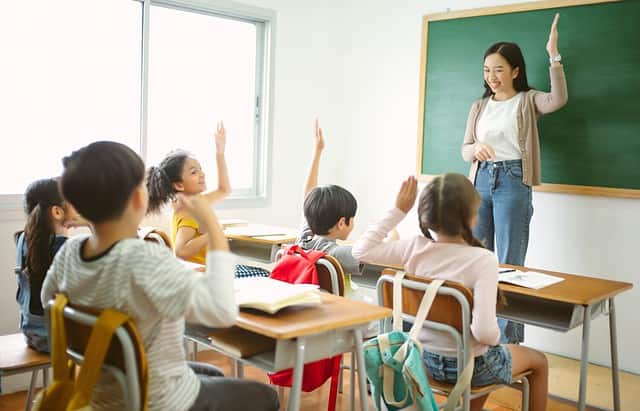 Group of Multi-ethnic happy elementary school children raising their arms to answer asian teacher question in classroom. Education, elementary school, learning, Back to school concept