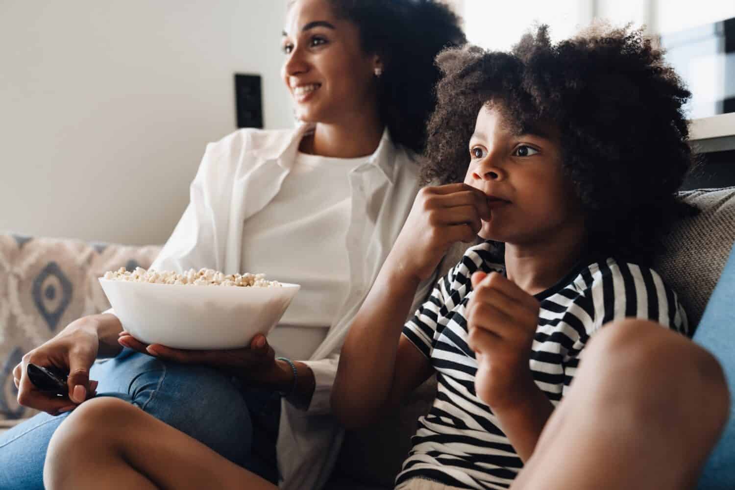 African american mom and son in casual wear eating popcorn and watching TV while sitting on sofa at home