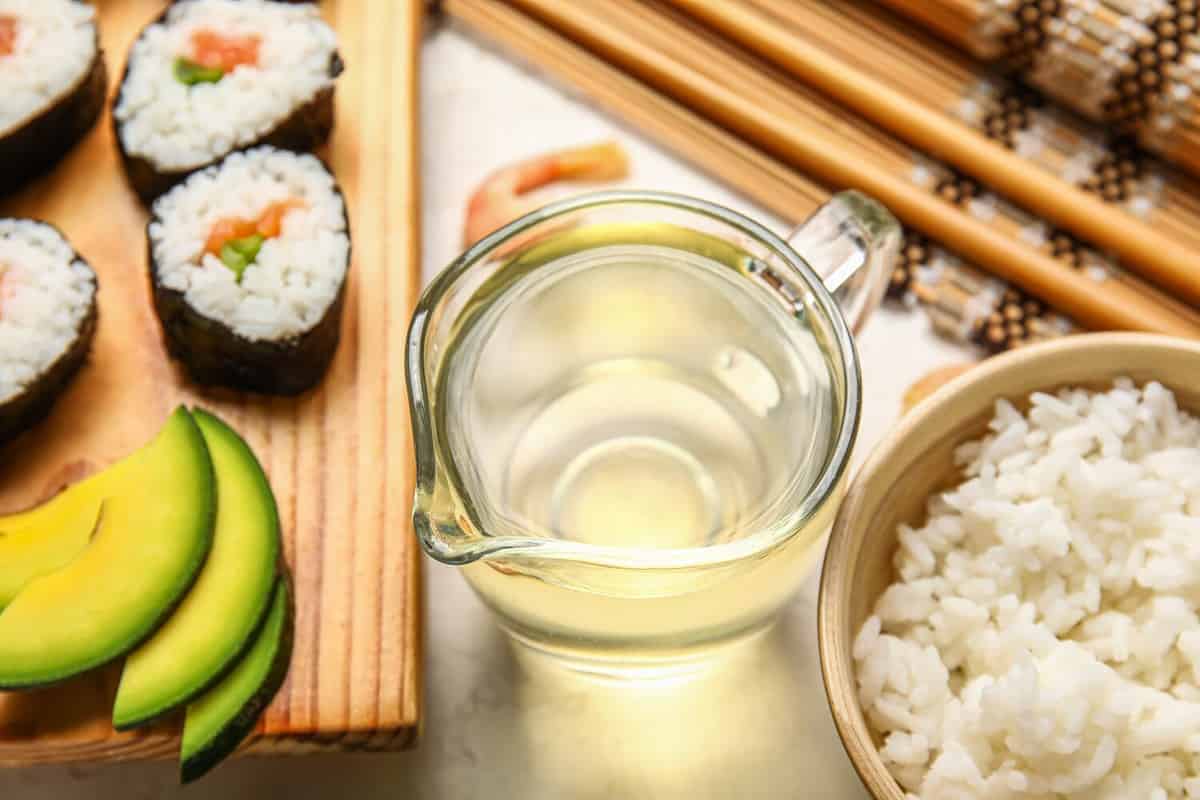 Jug with vinegar, boiled rice, avocado and sushi rolls on light table, closeup