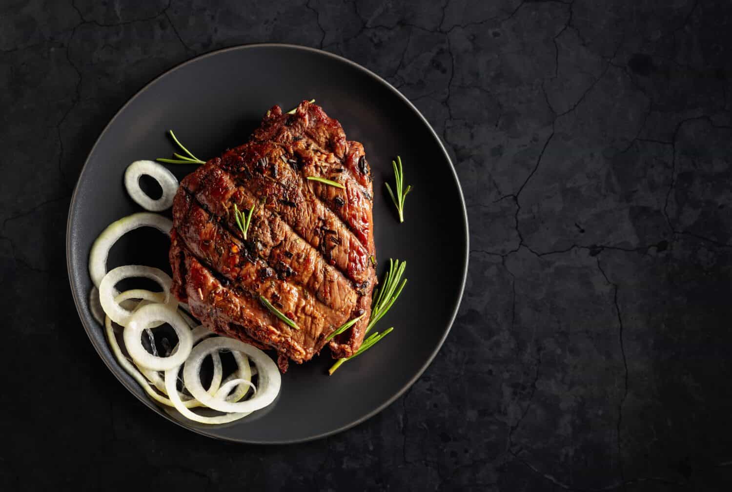 Grilled ribeye beef steak with rosemary and marinated onion on a black stone table. Copy space.