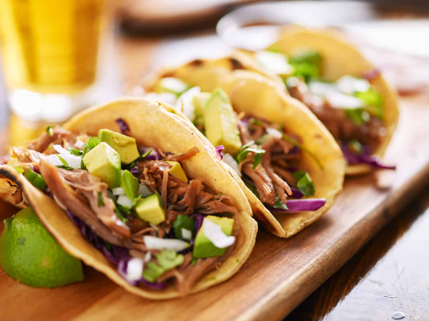 Tostadas vs. Tacos: three  pork carnitas street tacos in yellow corn tortilla with avocado, onion, cilantro and cabbage