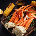 snow Crab legs served with melted butter, garlic cloves, lemon slices, grilled corn in cobs and fresh parsley on wooden cutting boards, horizontal view from above, close-up