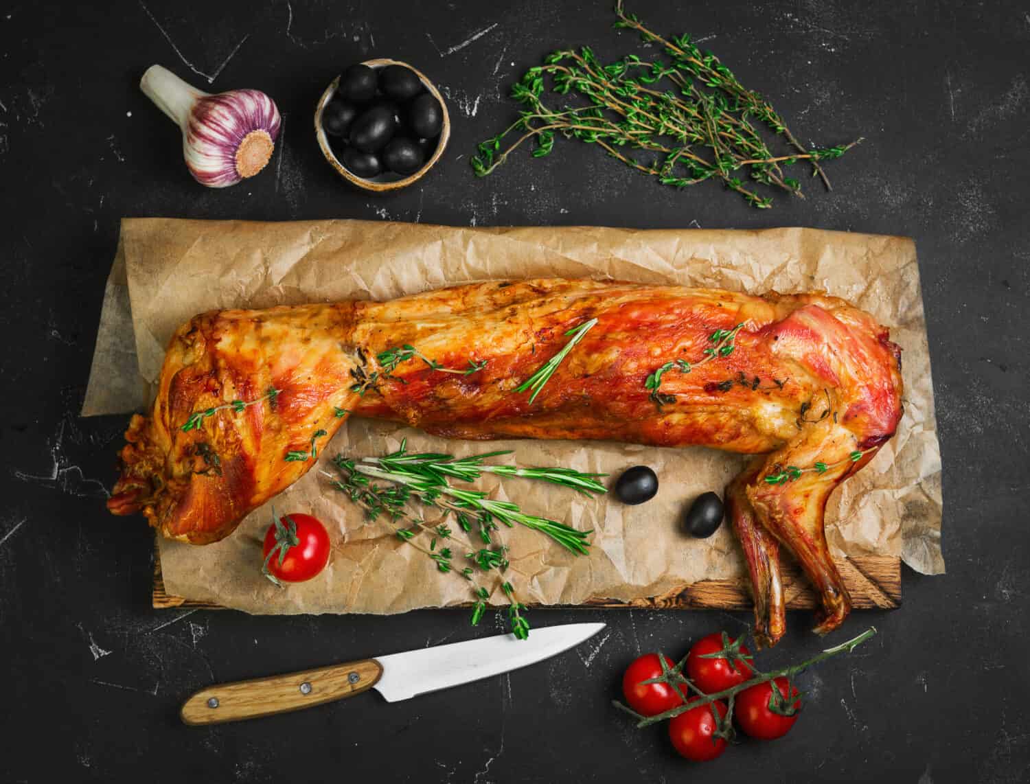 Roast baked whole rabbit on wooden board on paper. Ingredients for baked rabbit rosemary, thyme, cherry tomatoes, garlic, olives on dark background. A festive meal. Top view.  