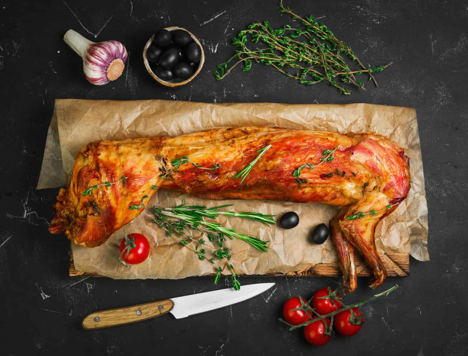 Roast baked whole rabbit on wooden board on paper. Ingredients for baked rabbit rosemary, thyme, cherry tomatoes, garlic, olives on dark background. A festive meal. Top view.