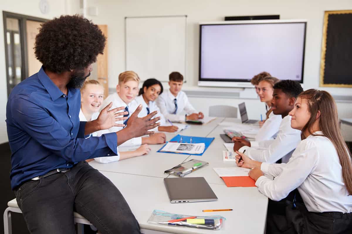 Male High School Teacher Sitting At Table With Teenage Pupils Wearing Uniform Teaching Lesson