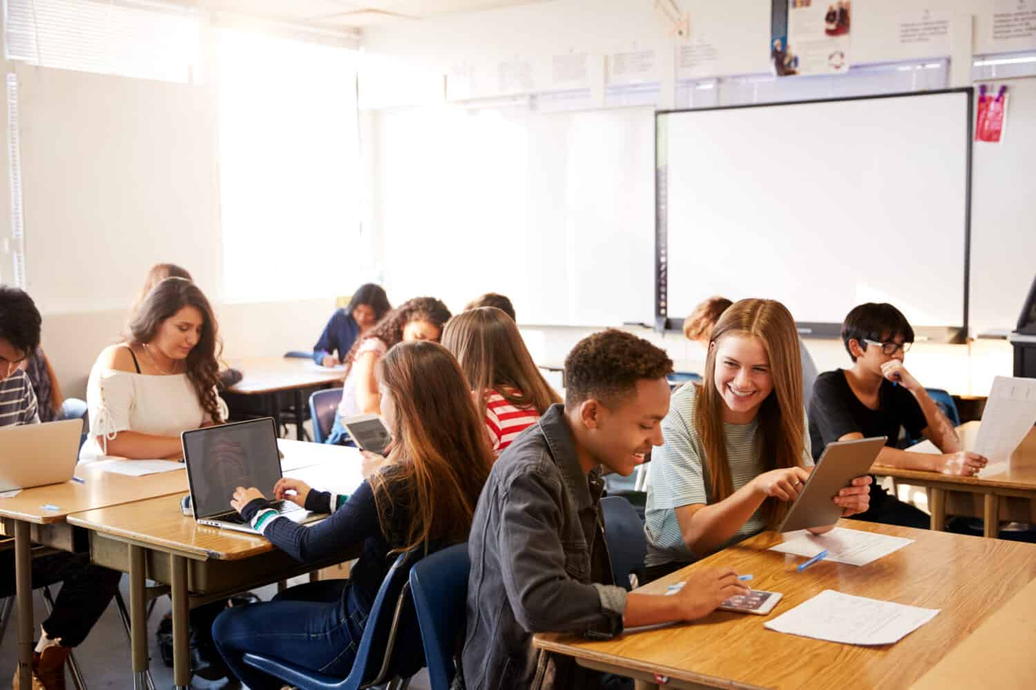 Wide Angle View Of High School Students Sitting At Desks In Classroom Using Laptops