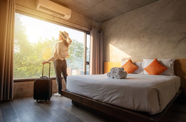 Portrait of tourist woman standing nearly window, looking to beautiful view with her luggage in hotel bedroom after check-in. Conceptual of travel and vacation.