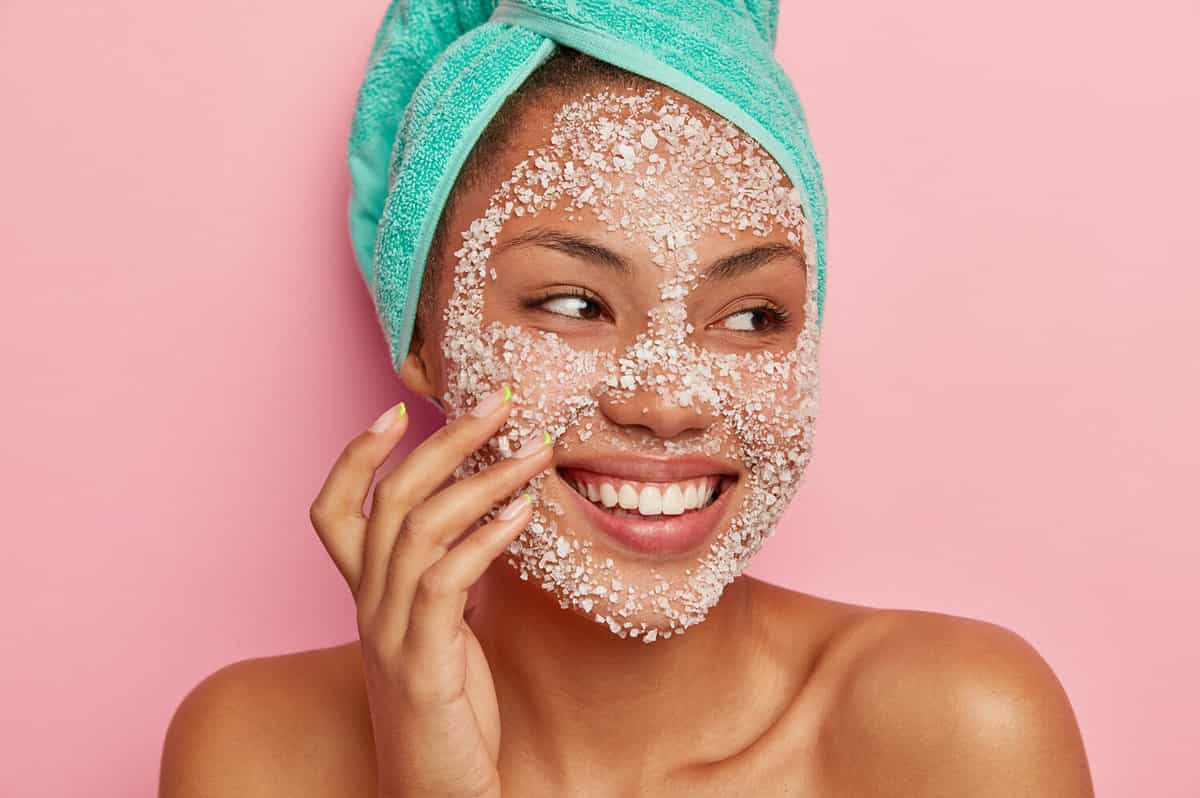 Close up portrait of glad African American woman massages cheeks, applies sea salt scrub, looks away, has gentle smile, shows white teeth, wears turquoise towel, happy to get spa treatments.