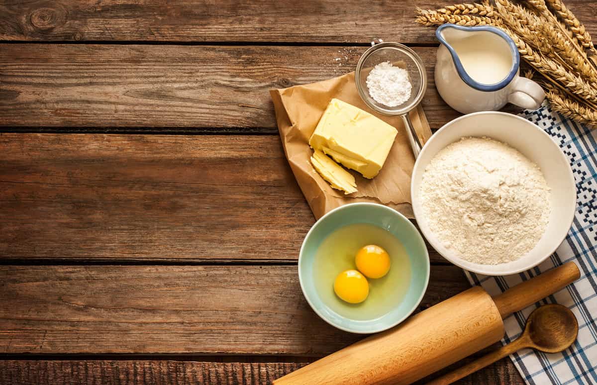 Baking cake in rural kitchen - dough recipe ingredients (eggs, flour, milk, butter, sugar) and rolling pin on vintage wood table from above. Rustic background with free text space.