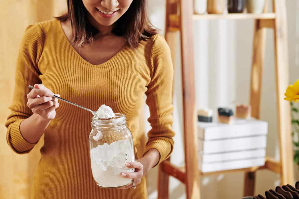 Smiling young woman taking spoon of soda lye from glass jar when making fragrant soap at home