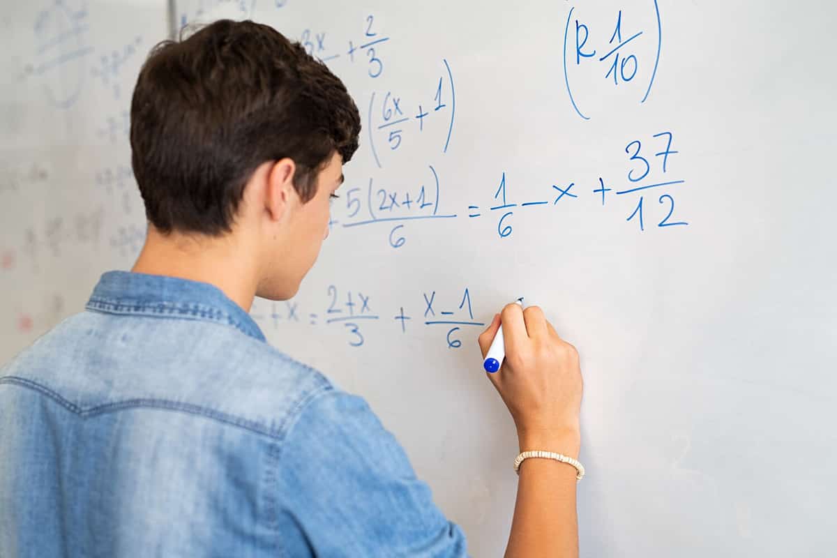 Back view of high school student solving math problem on whiteboard in classroom. Young man writing math solution on white board using marker. College guy solving math expression during lesson.