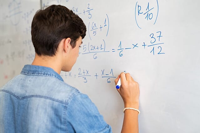 Back view of high school student solving math problem on whiteboard in classroom. Young man writing math solution on white board using marker. College guy solving math expression during lesson.