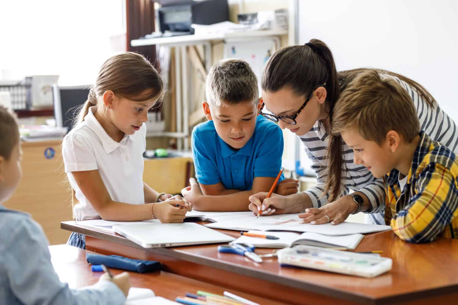 Female teacher helps school kids to finish they lesson.They sitting all together at one desk.	