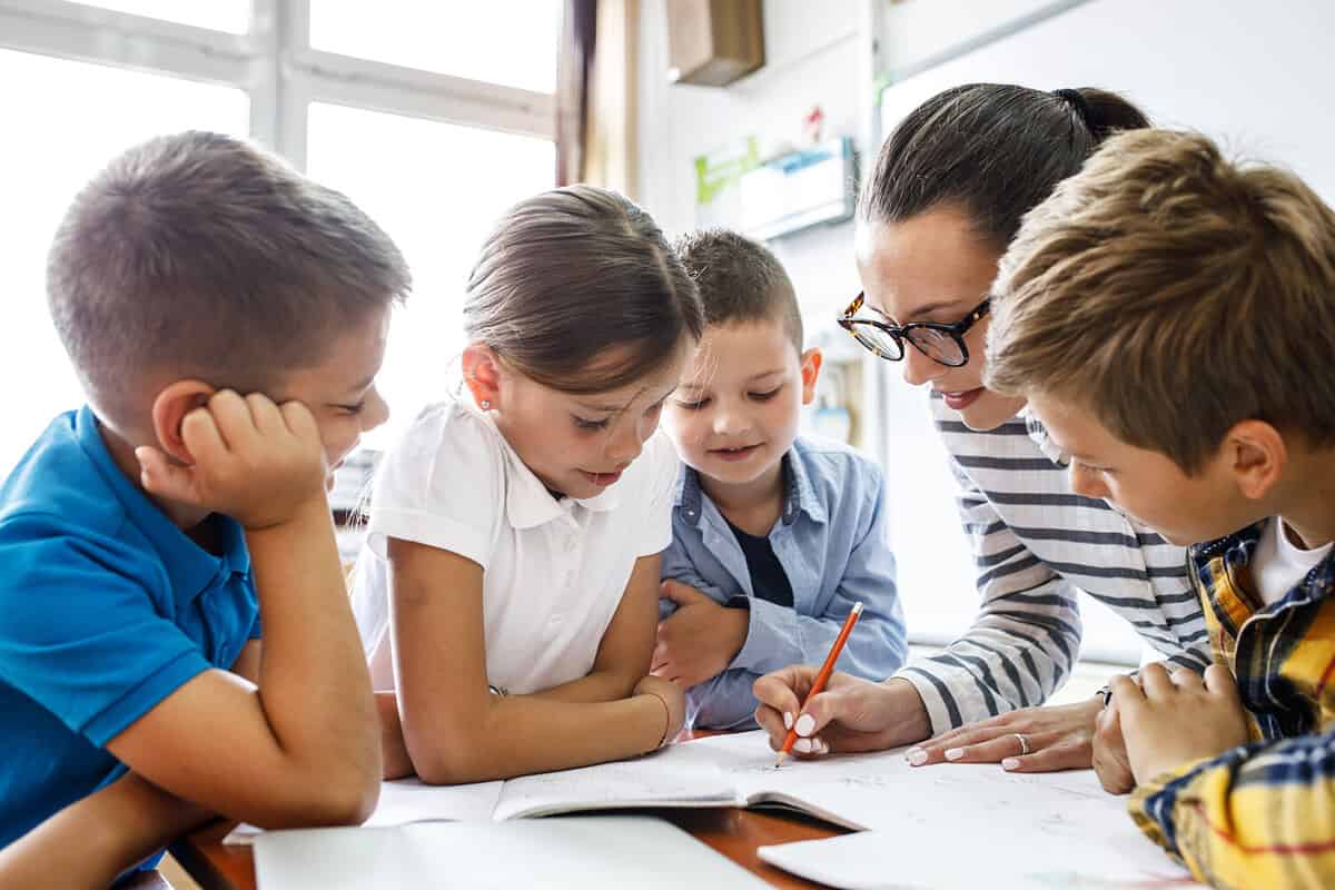 Female teacher helps school kids to finish they lesson.They sitting all together at one desk.