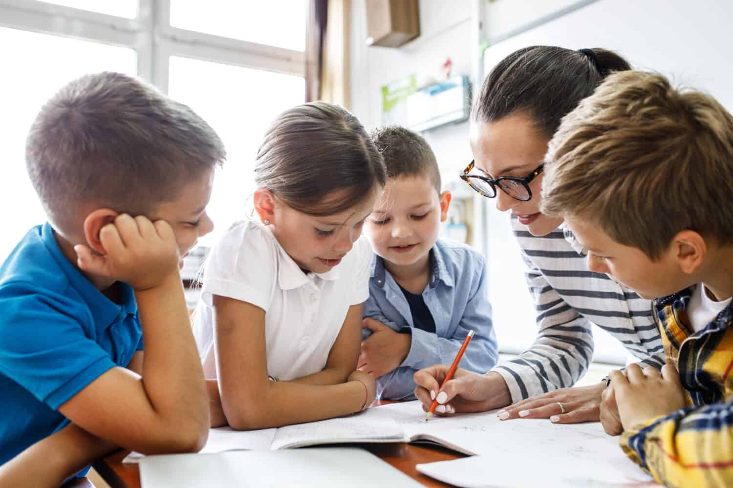 Female teacher helps school kids to finish they lesson.They sitting all together at one desk.	