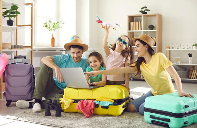 Happy excited family and daughter children buying payment for flight ticket booking hotel online using laptop with travel suitcase around at home living room. Holiday vacation traveling abroad concept