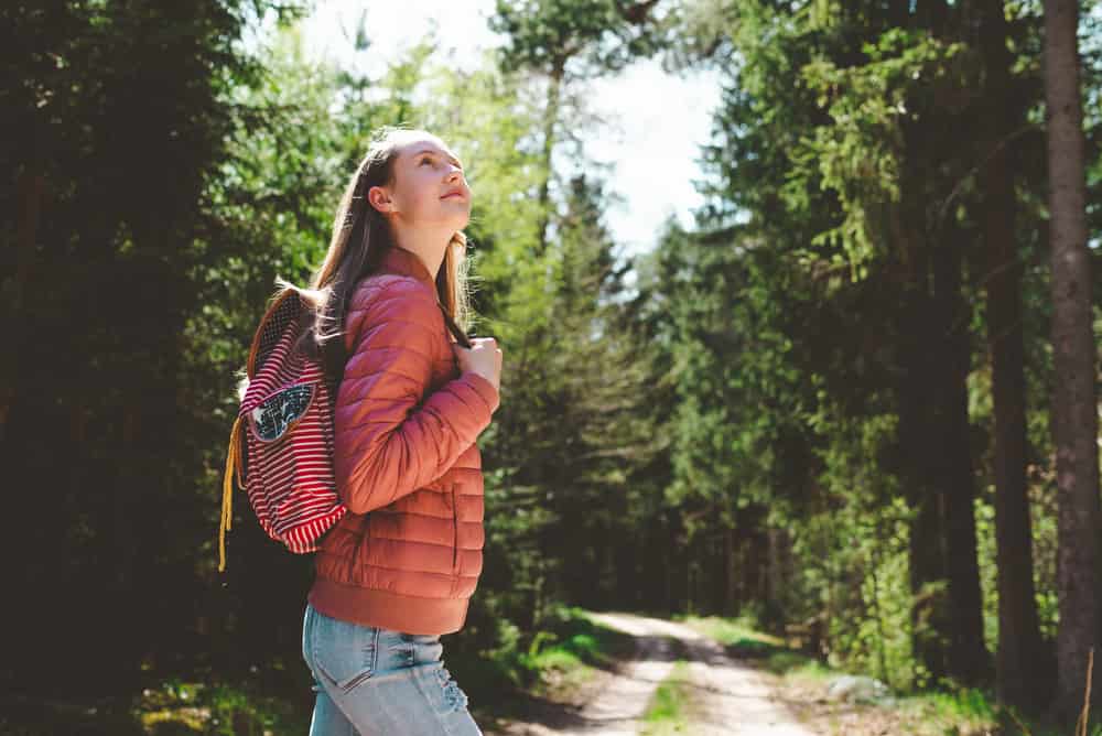 Teen tourist girl admiring forest, red puffer jacket and orange hiking backpack.