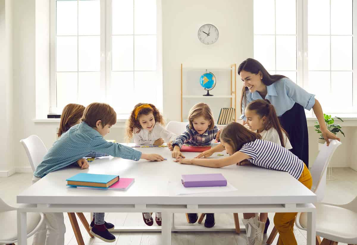 Happy school teacher and first grade students having interesting engaging activities in class. Little children sitting around big classroom table, learning ABCs, talking and playing fun games together