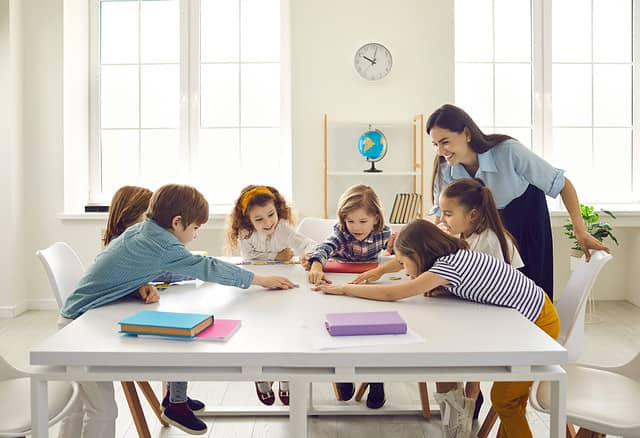 Happy school teacher and first grade students having interesting engaging activities in class. Little children sitting around big classroom table, learning ABCs, talking and playing fun games together