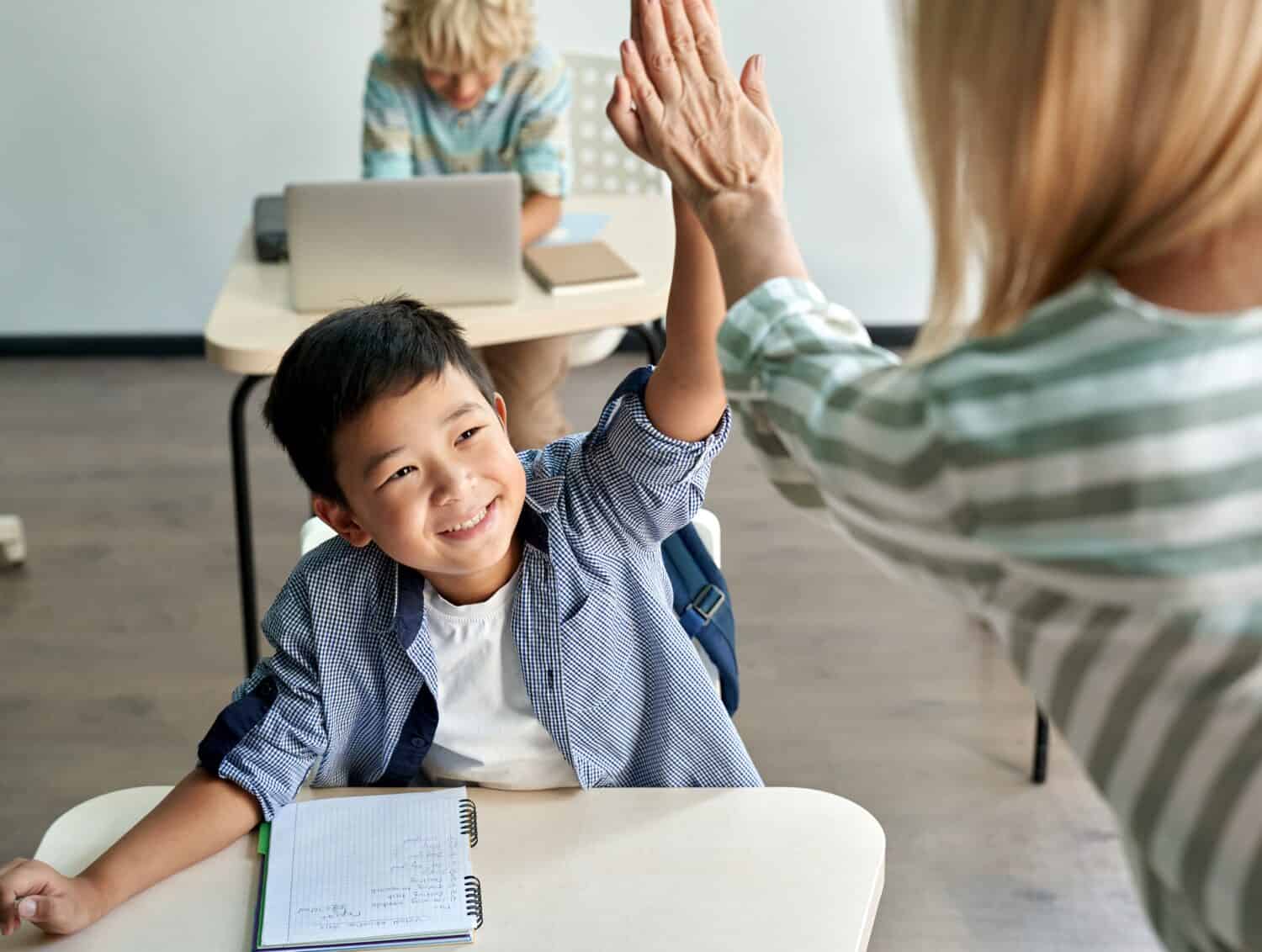 Happy Asian kid boy giving high five to female teacher at class in classroom. Teacher encouraging cheerful chinese helping child student giving support during elementary junior school lesson.