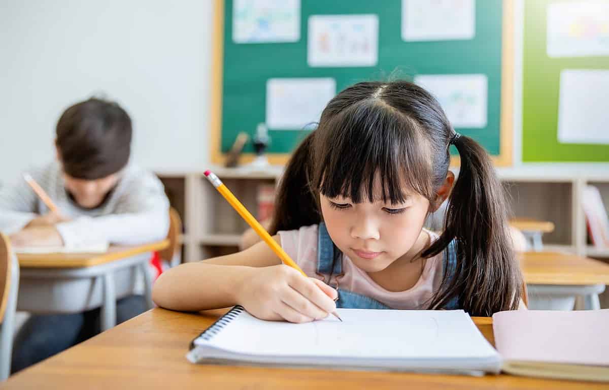 Portrait of little pupil writing at desk in classroom at the elementary school. Student girl study doing test in primary school. Children writing notes in classroom. Education knowledge concept