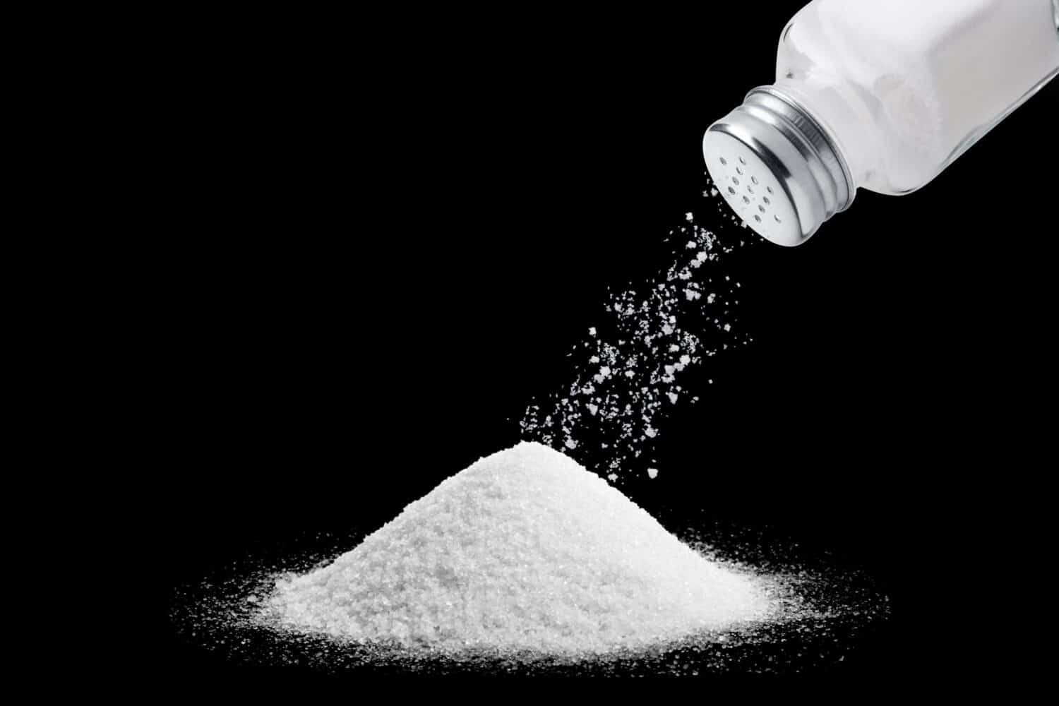 image of salt powder piled on a black background in the upper right corner a bottle of salt sprinkled with salt powder out of the bottle onto the pile of salt below. Suitable for use in food media.
