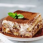 Perfect homemade tiramisu cake with fresh mint. Tiramisu portion on pink plate over white marble background. Delicious no bake tiramisu in natural daylight. Close up