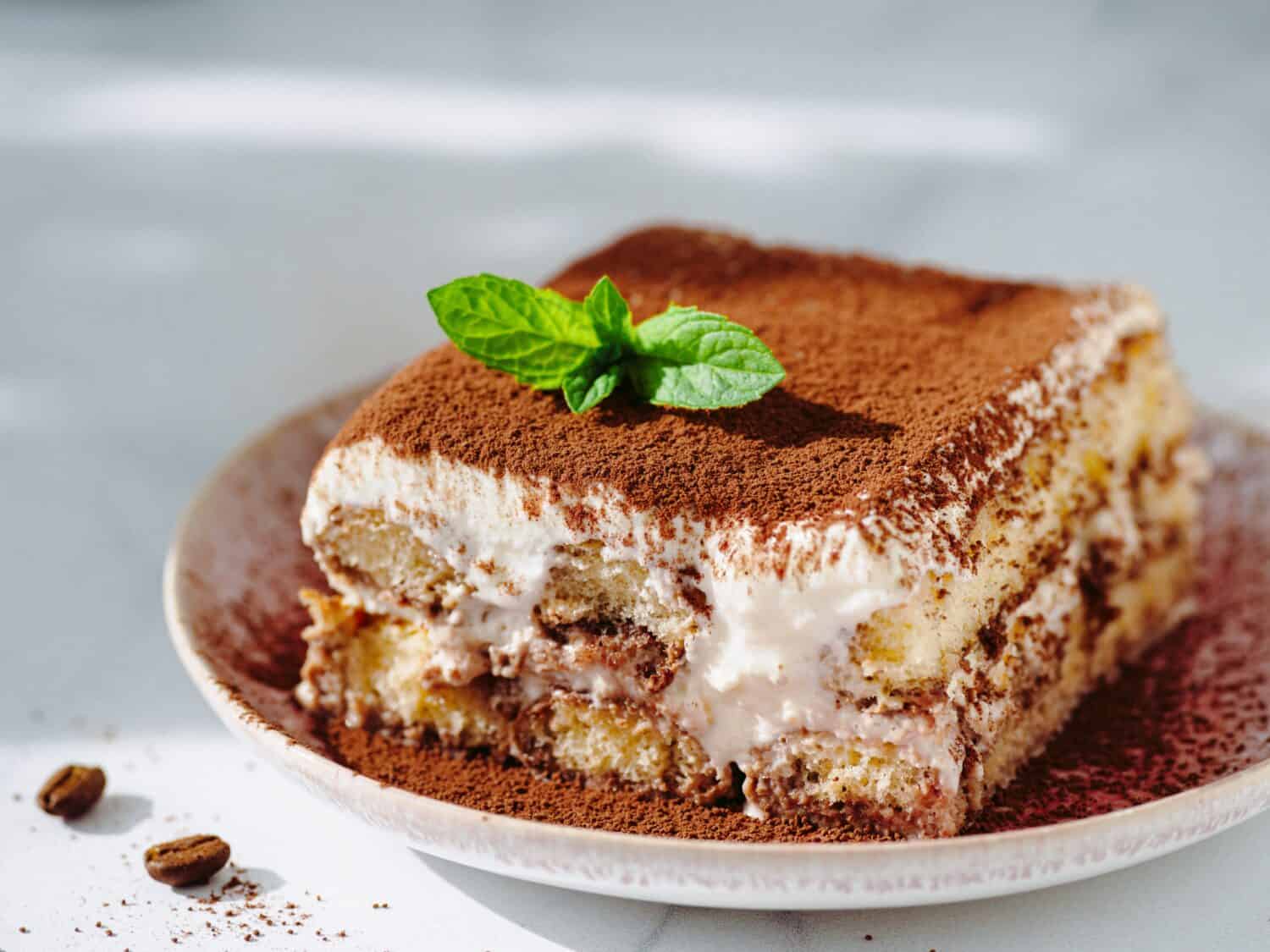 Perfect homemade tiramisu cake with fresh mint. Tiramisu portion on pink plate over white marble background. Delicious no bake tiramisu in natural daylight. Close up