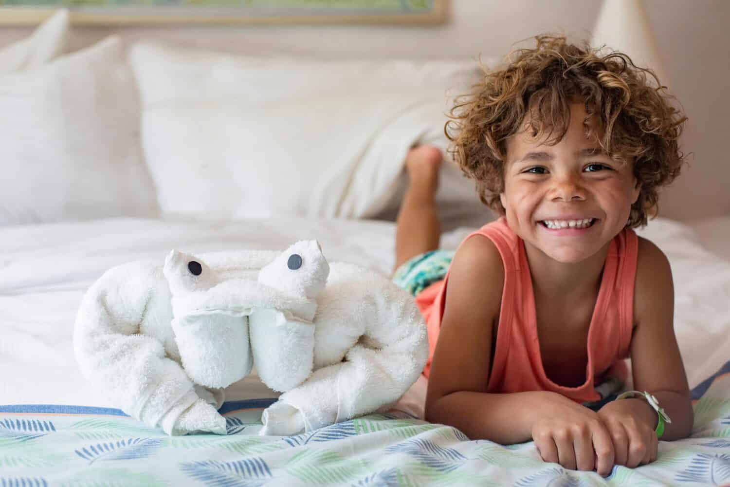 Beautiful young diverse boy sitting on bed in a cruise ship next to a towel animal. Enjoying a fun cruise vacation in his luxury cruise ship cabin with his family
