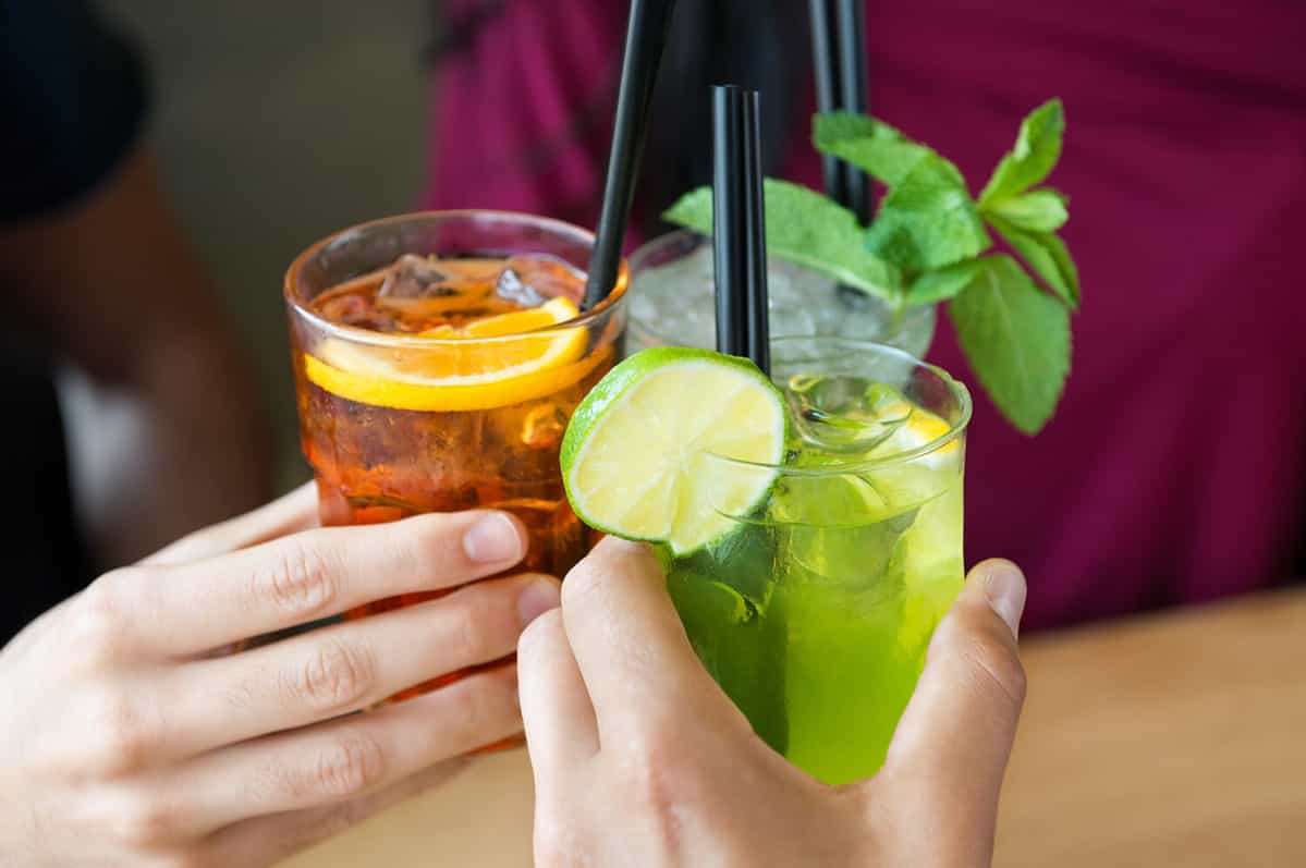 Closeup shot of friends toasting with cocktails. Young people drinking at aperitif. Shallow depth of field with focus on friends hand toasting juice glass. Close up of hands holding a cocktail glass.