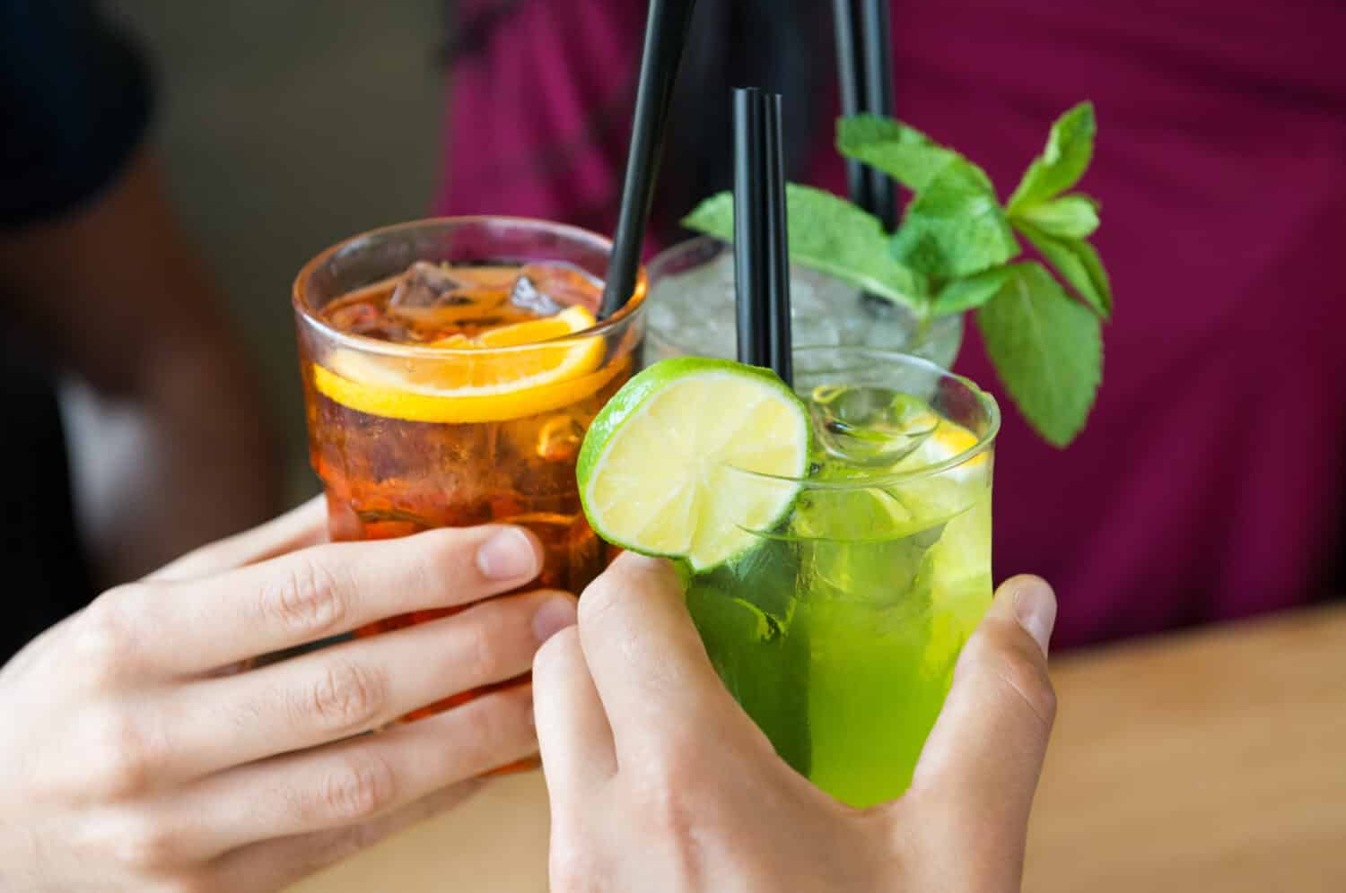 Closeup shot of friends toasting with cocktails. Young people drinking at aperitif. Shallow depth of field with focus on friends hand toasting juice glass. Close up of hands holding a cocktail glass.