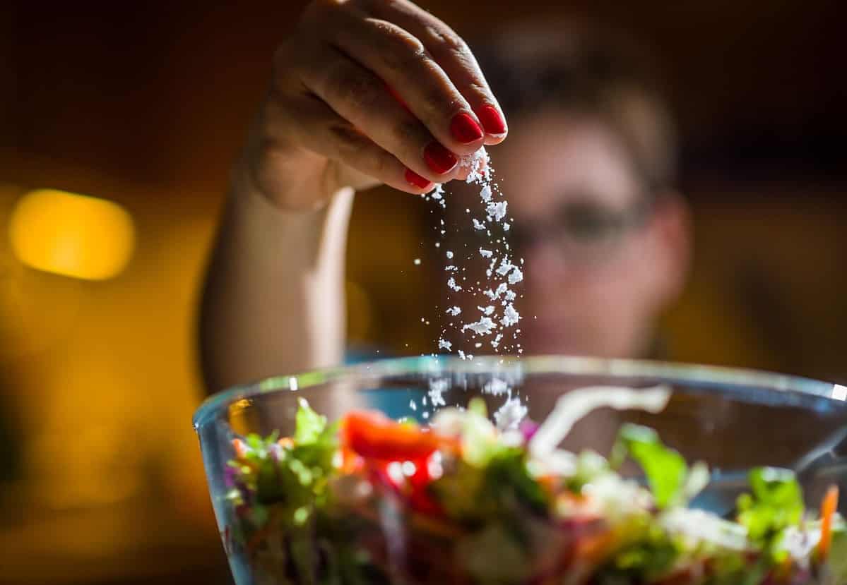 Woman preparing healthy salad in kitchen, adding salt to the bowl.