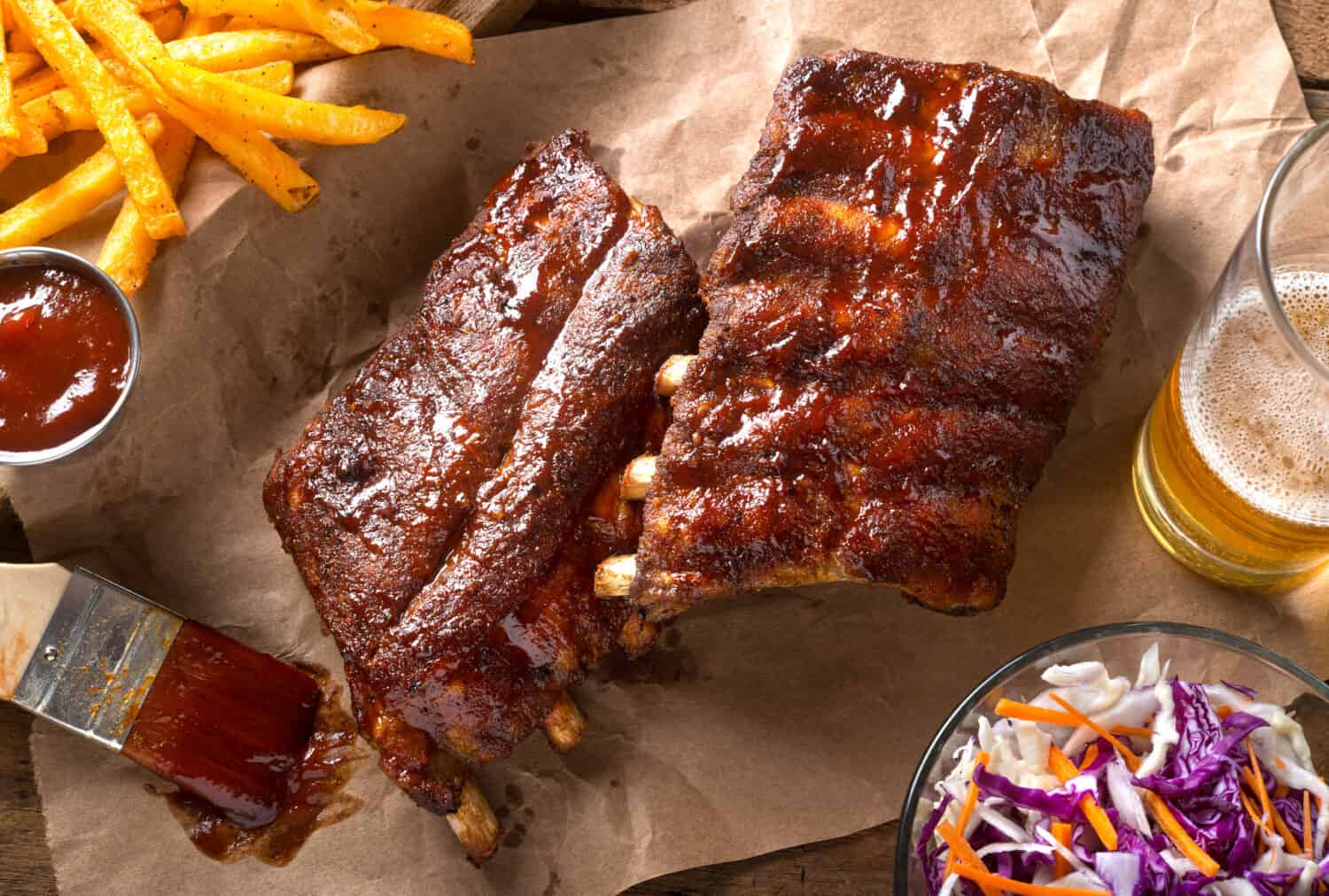 A rack of delicious baby back ribs with barbecue sauce, french fries, coleslaw and beer.