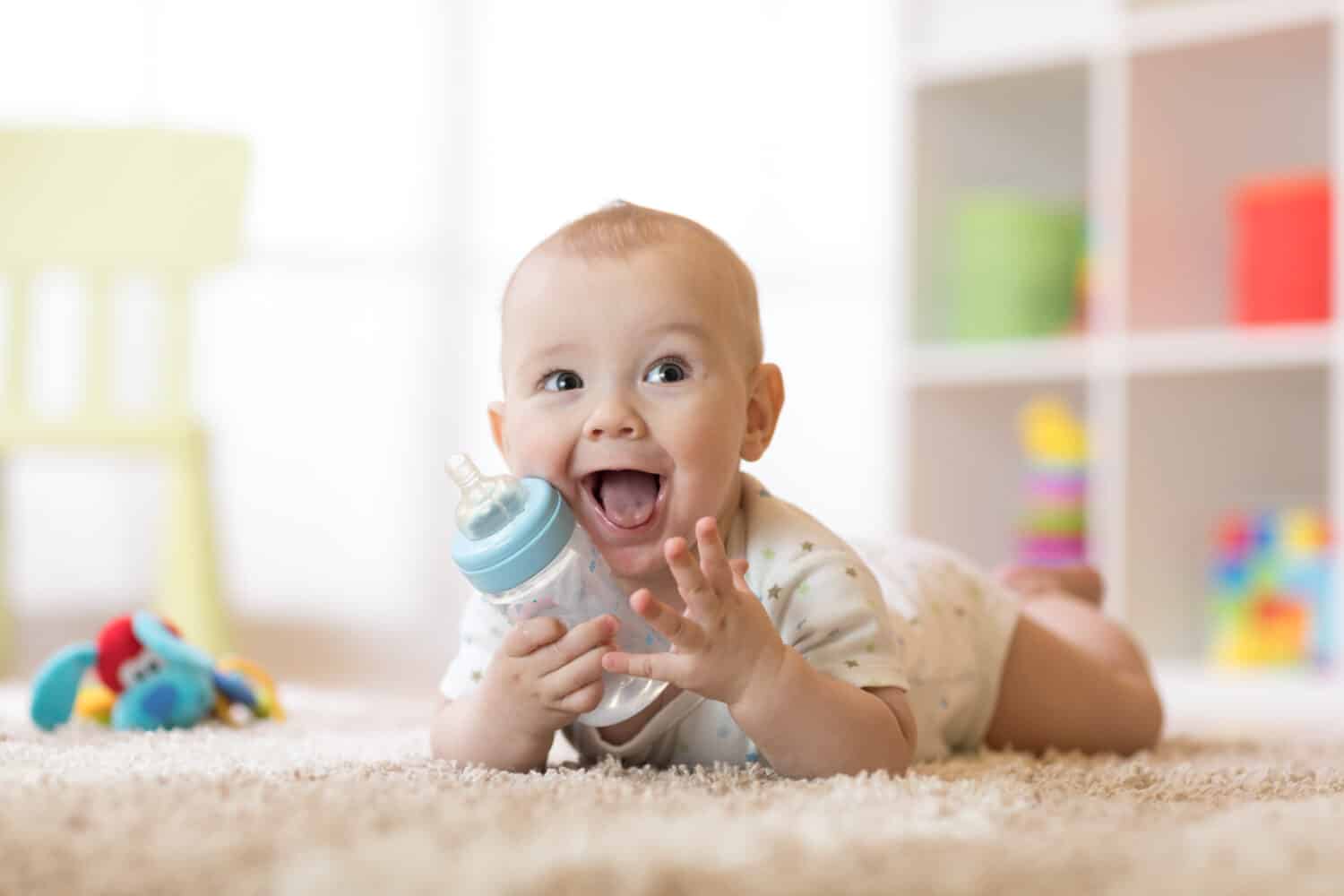 Cute baby boy drinking from bottle. Kid lying on carpet in nursery at home. Smiling child is 7 months old.