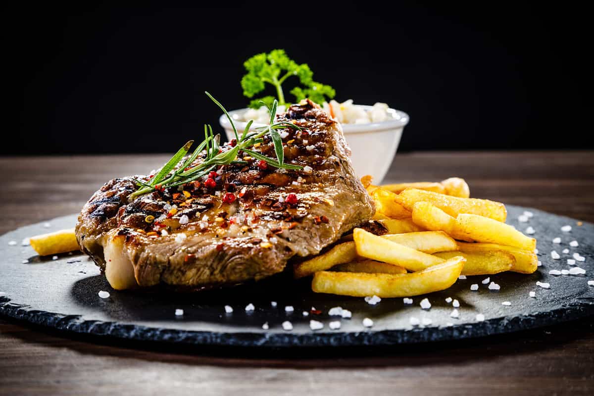 Grilled steak with french fries and vegetables served on black stone on wooden table