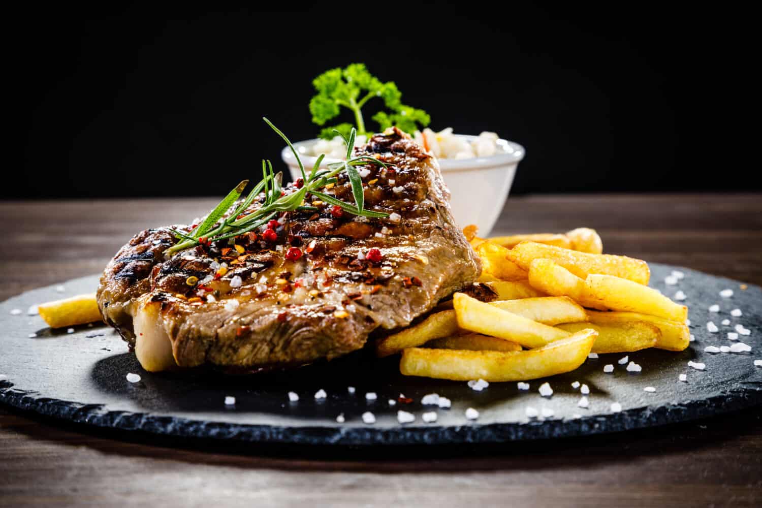 Grilled steak with french fries and vegetables served on black stone on wooden table 