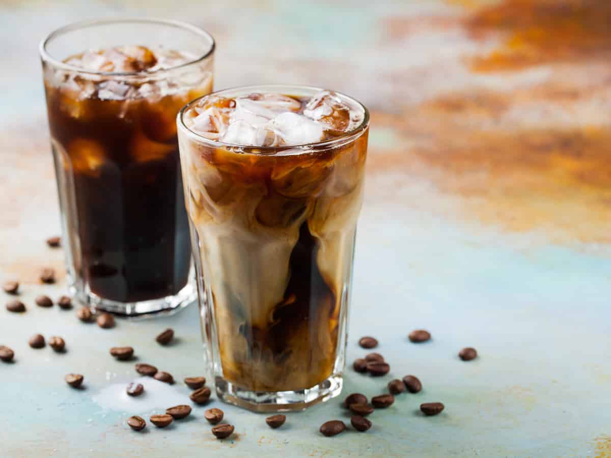 Ice coffee in a tall glass with cream poured over and coffee beans. Cold summer drink on a blue rusty background with copy space